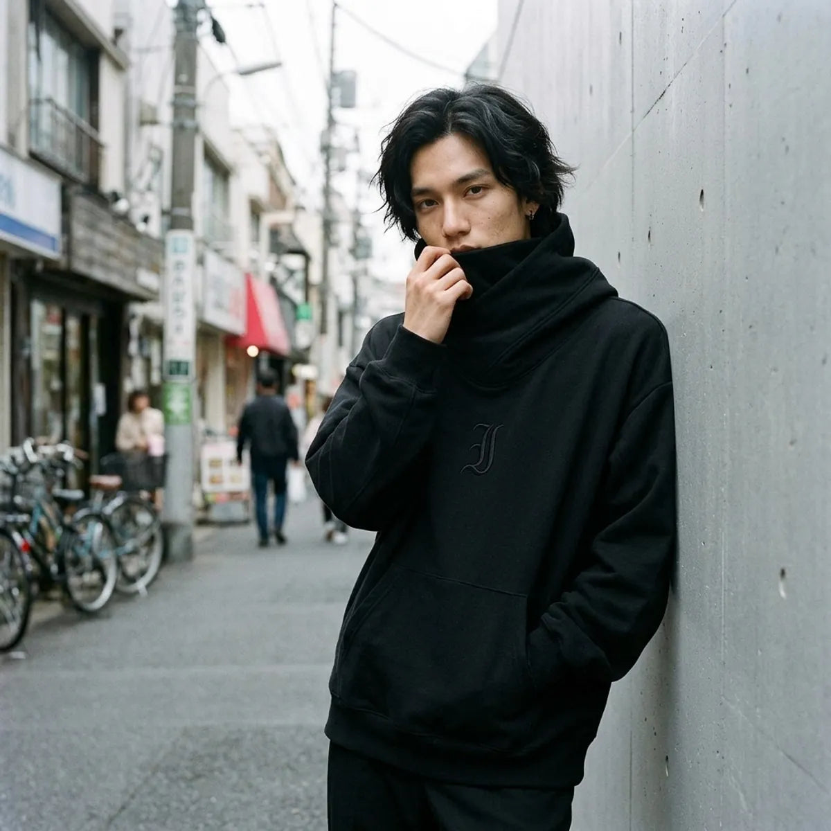 Schwarzer Streetwear-Hoodie aus Tokio, getragen mit hochgezogenem Kragen über dem unteren Gesicht vor einer Wand
